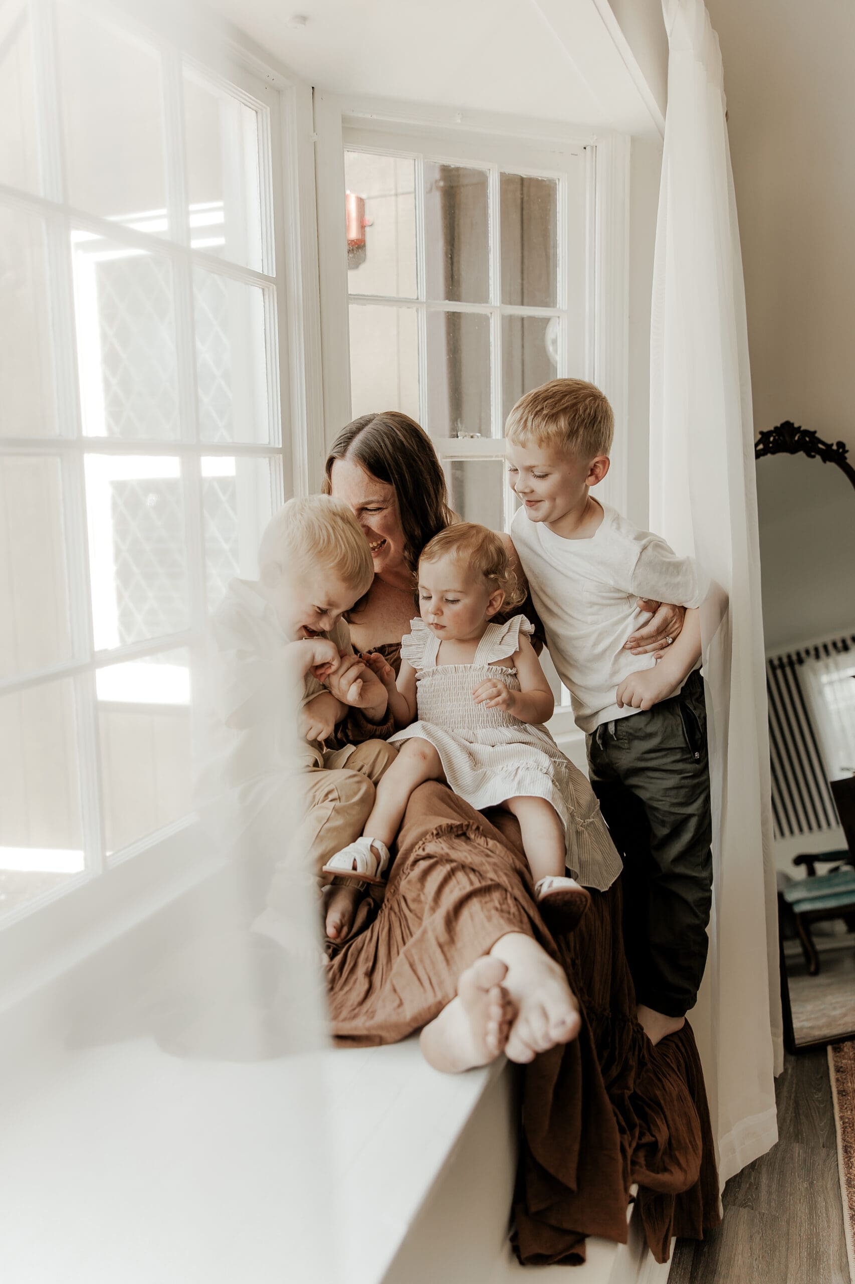 Mother sitting on a window seat with her three young children in a cozy, naturally lit photography studio in Mendham, NJ. Soft light filters through large windows as the family shares a tender, candid moment.