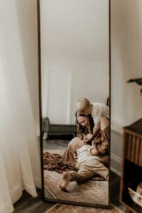 Candid reflection of a mother playing and laughing with her two young sons in a warm, neutral-toned photography studio. Captured in a floor-length mirror with natural light and soft furnishings visible in the background.