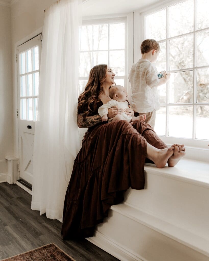 Barefoot mother sits with her baby and young son by a large window in a light-filled photography studio in Mendham, NJ, capturing relaxed family moments in a bedroom-inspired setting.