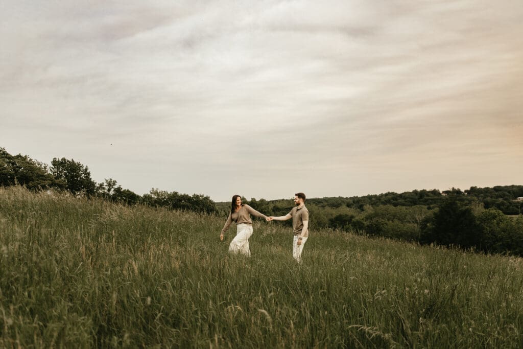 Couple holding hands and walking through a tall grassy field during golden hour; candid outdoor photography session in the rolling hills of Far Hills, New Jersey.