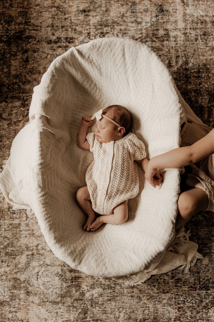Newborn baby lying peacefully in a soft, knit romper on a cozy lounger with sibling’s hand resting gently nearby — lifestyle newborn session in Budd Lake, New Jersey.
