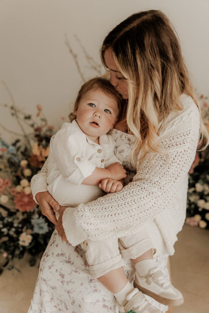 Toddler looking at the camera while being held in mother’s arms, surrounded by soft floral tones — motherhood session filled with tenderness and connection in Princeton, NJ.