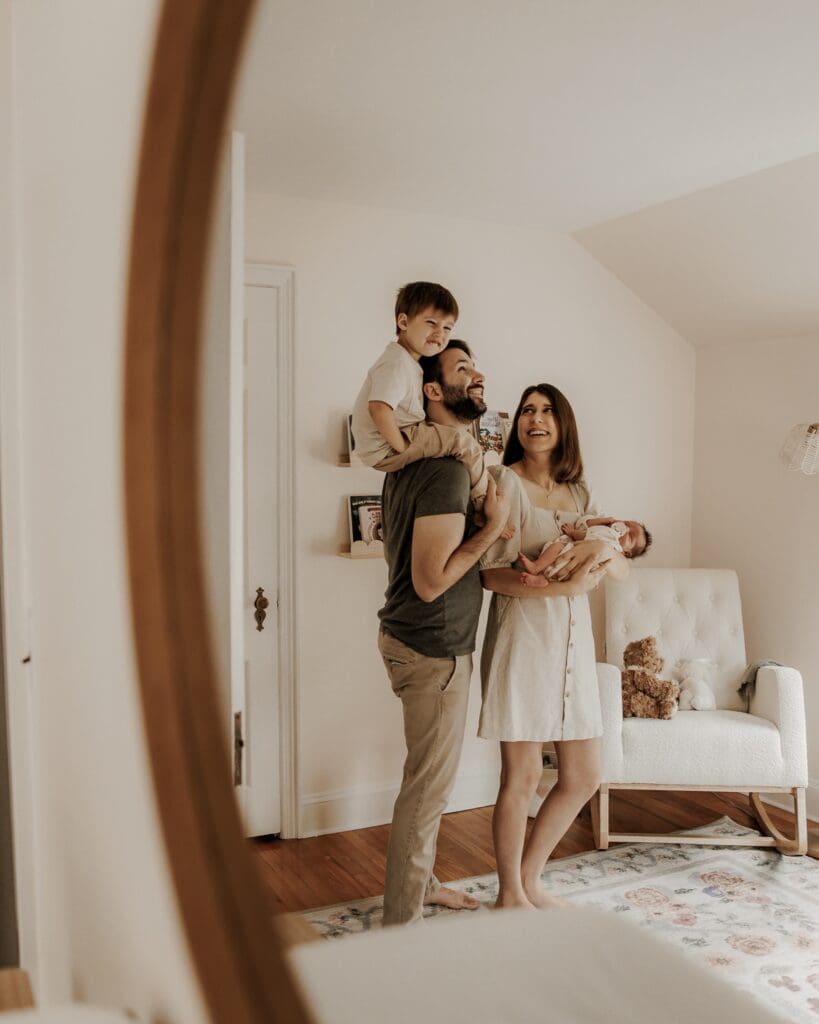 Joyful family of four reflected in a mirror, with dad holding toddler on his shoulders and mom cradling a newborn — warm in-home family session in Morristown, NJ.