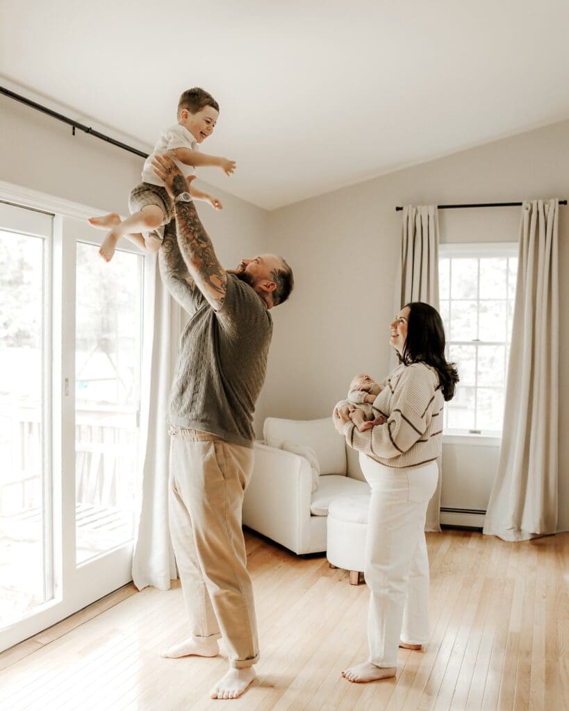 Father playfully lifting toddler near a bright window as mother smiles while holding newborn — lifestyle newborn and family session in a sun-filled home in Basking Ridge, NJ.
