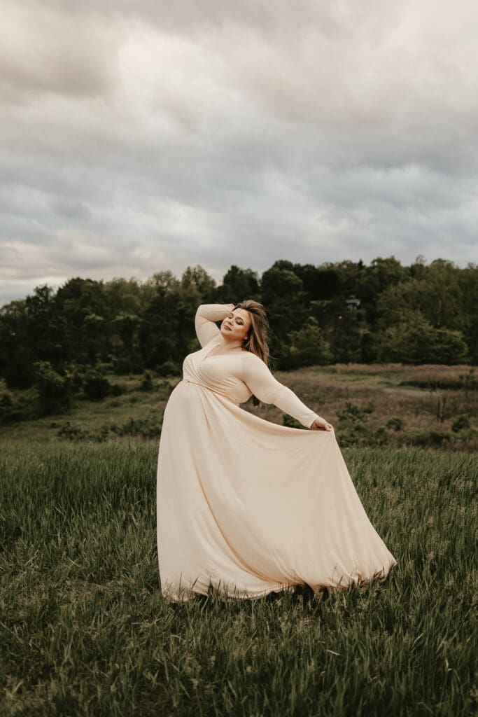 Expecting mother in a flowing blush gown striking a confident pose in an open meadow under moody skies — elegant maternity photo session in Far Hills, NJ.