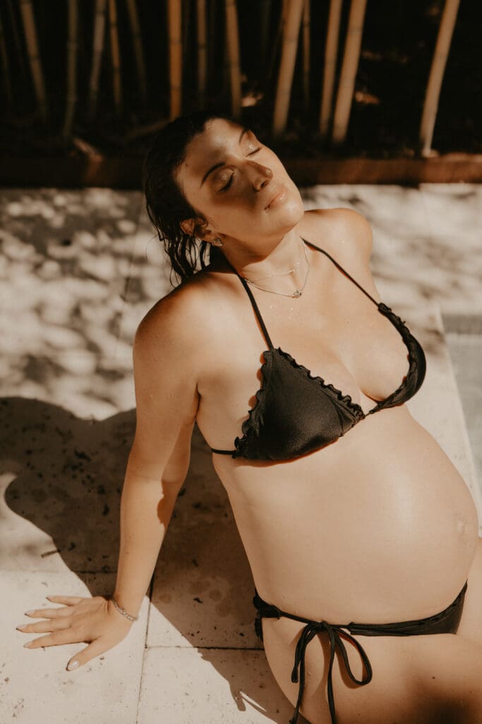 Pregnant woman in black bikini soaking in the sun by a poolside, embracing the beauty of motherhood during a warm, golden maternity session. Located in Asbury Park, NJ.