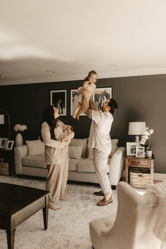 Father lifting toddler in the air while mother holds newborn nearby, all barefoot and cozy in their warmly lit living room — candid in-home family session in Rockaway, NJ.