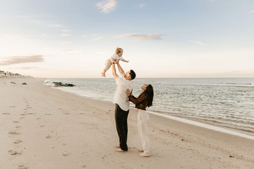 Father lifting baby into the air with ocean waves and soft sunset light in the background, as mother smiles beside them — joyful family session in Sea Bright, NJ.