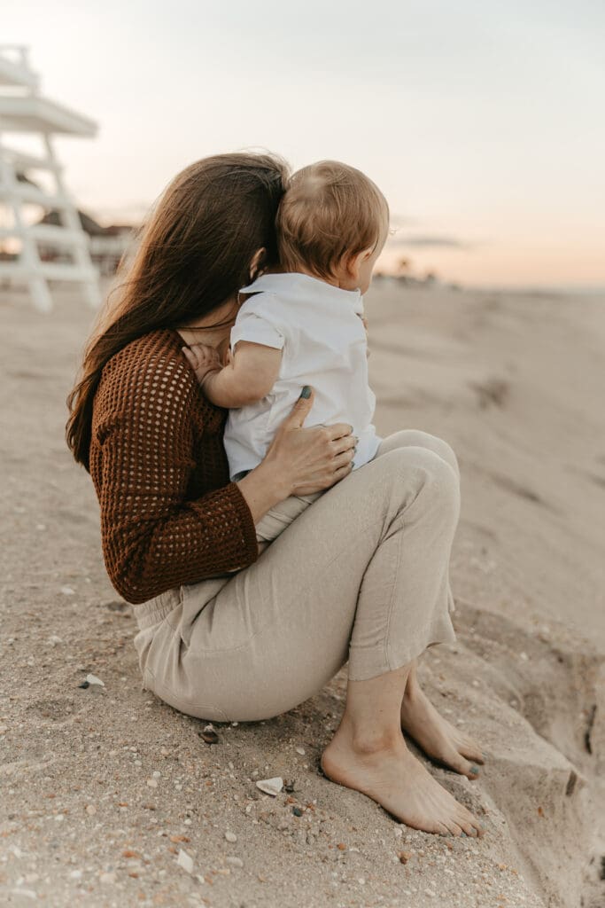Mother sitting barefoot in the sand, holding toddler close while watching the sunset at Sea Bright beach in New Jersey — natural and intimate motherhood photography.